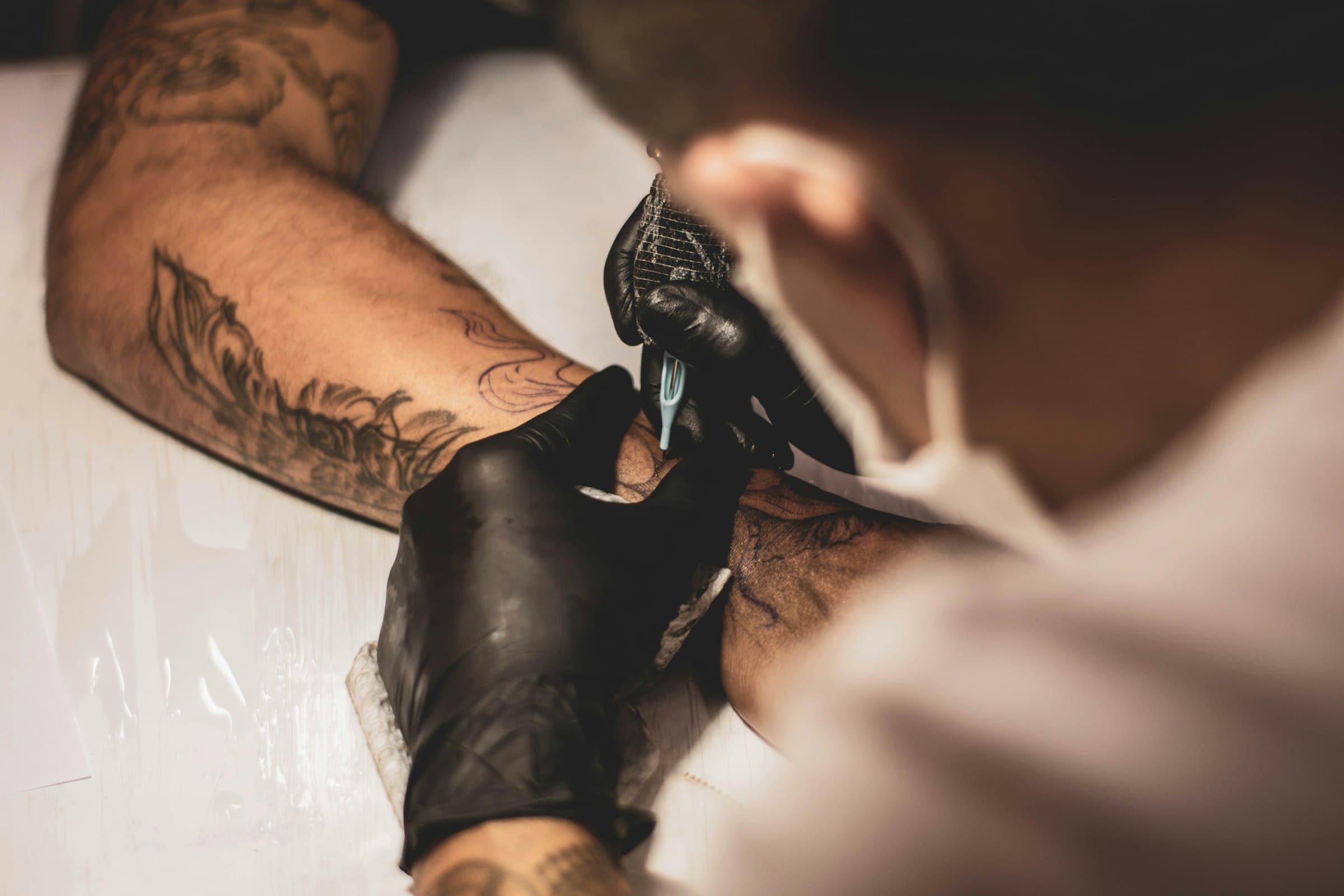 La main gantée d'un tatoueur, la machine posée sur l'avant-bras d'un client, encre et lumière chaude.